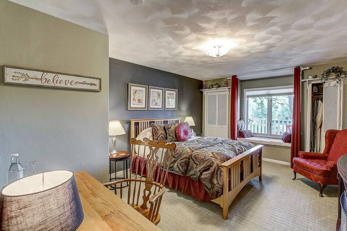 A bedroom with a bed, a chair, a bedside table, a lamp, and a window with curtains.