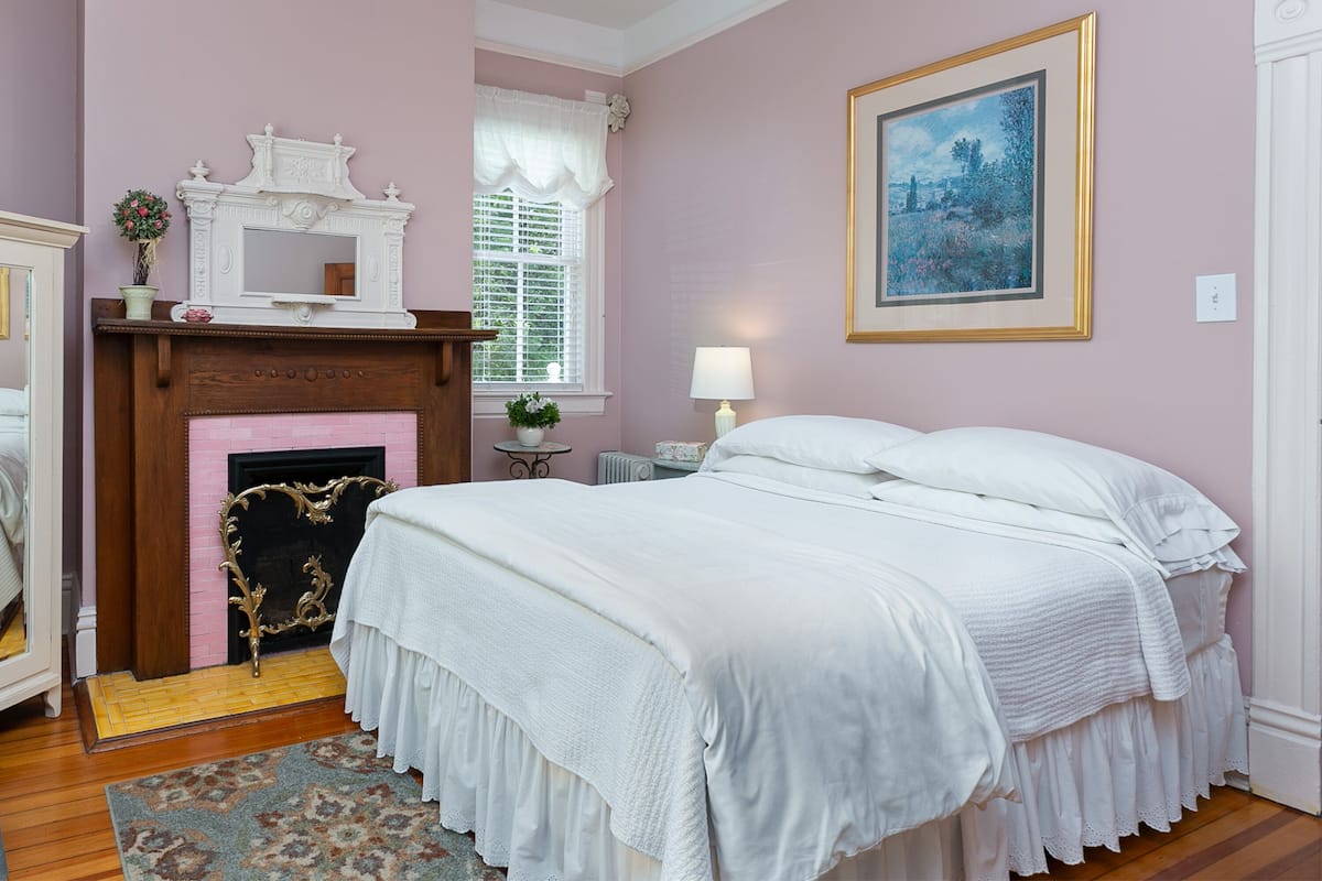 A bedroom with a large bed, a fireplace, a window with white shutters, and a framed painting on the wall.
