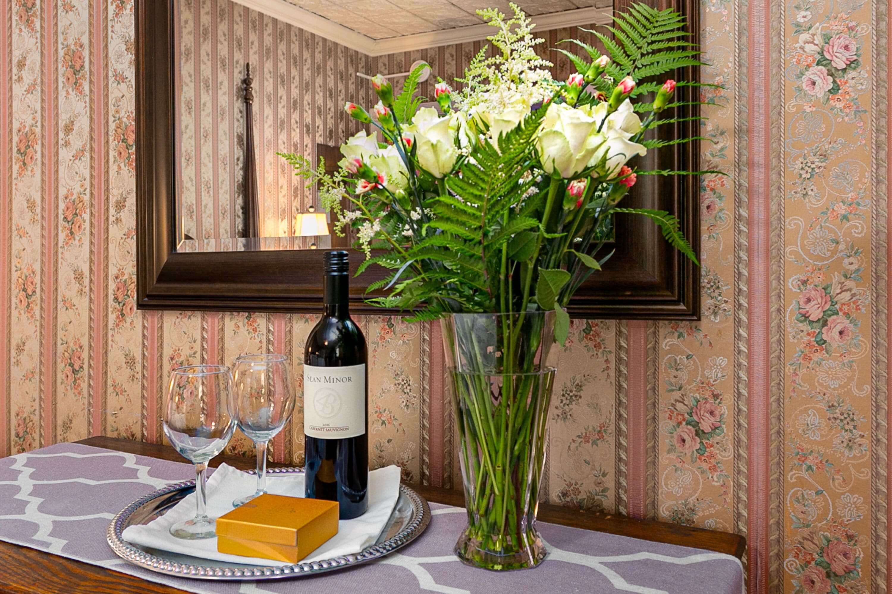 A table with a bottle of wine, two glasses, a tray with a box, and a vase with flowers.