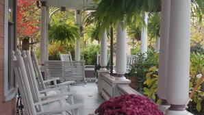 A covered porch with white chairs and a bench, surrounded by columns and potted plants. - Beaufort House Inn (Asheville)