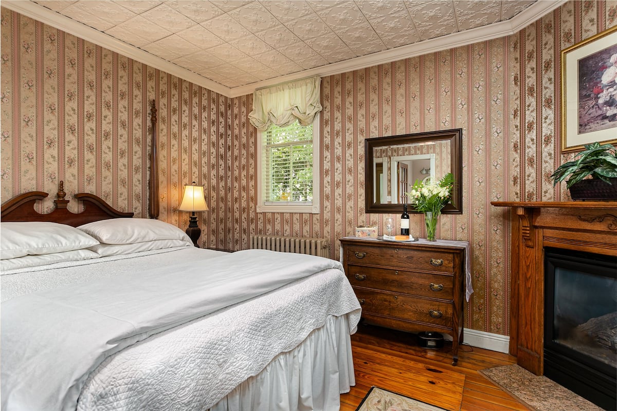 A bedroom with a large bed, a dresser, a mirror, a window with curtains, a fireplace, and a radiator.