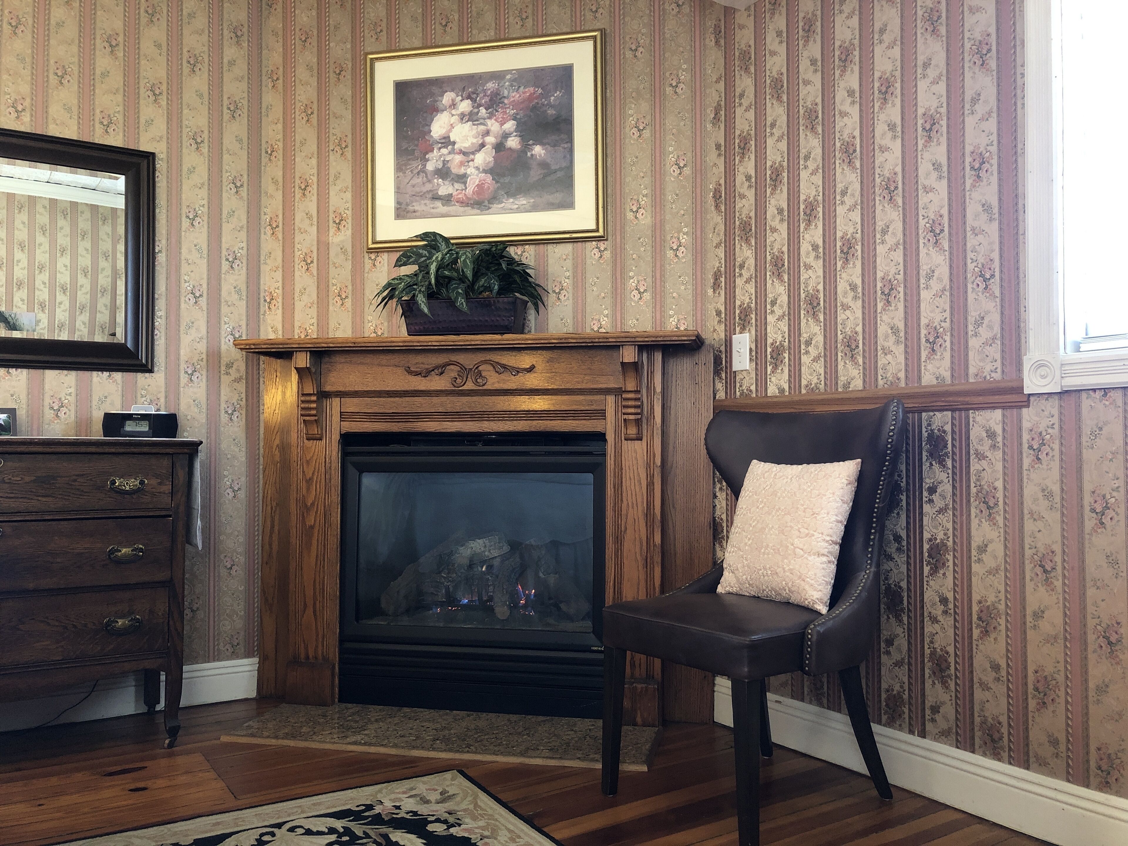 A room with a fireplace, a chair, a dresser, and a framed floral painting on the wall.