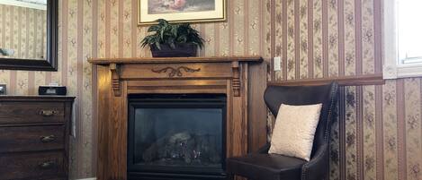 A room with a fireplace, a chair, a dresser, and a framed floral painting on the wall.