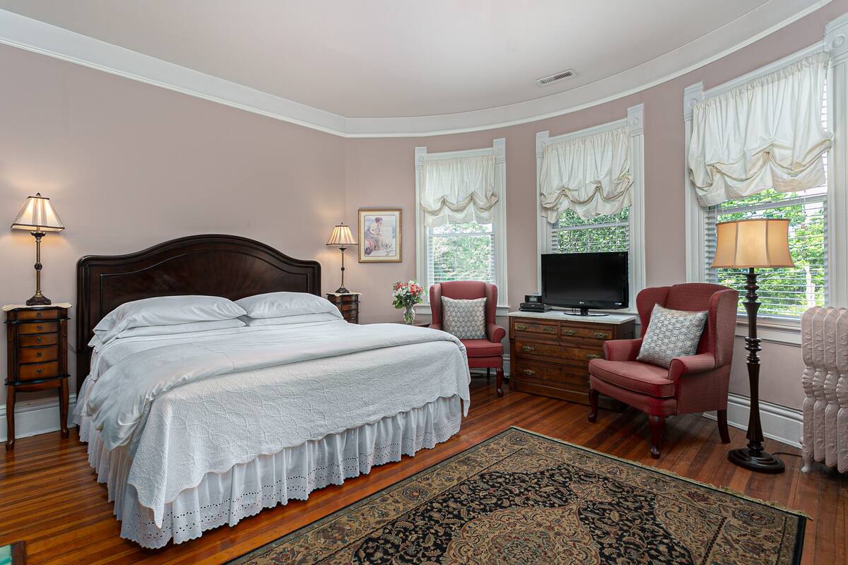 A bedroom with a large bed, a desk, a chair, a television, and two lamps.