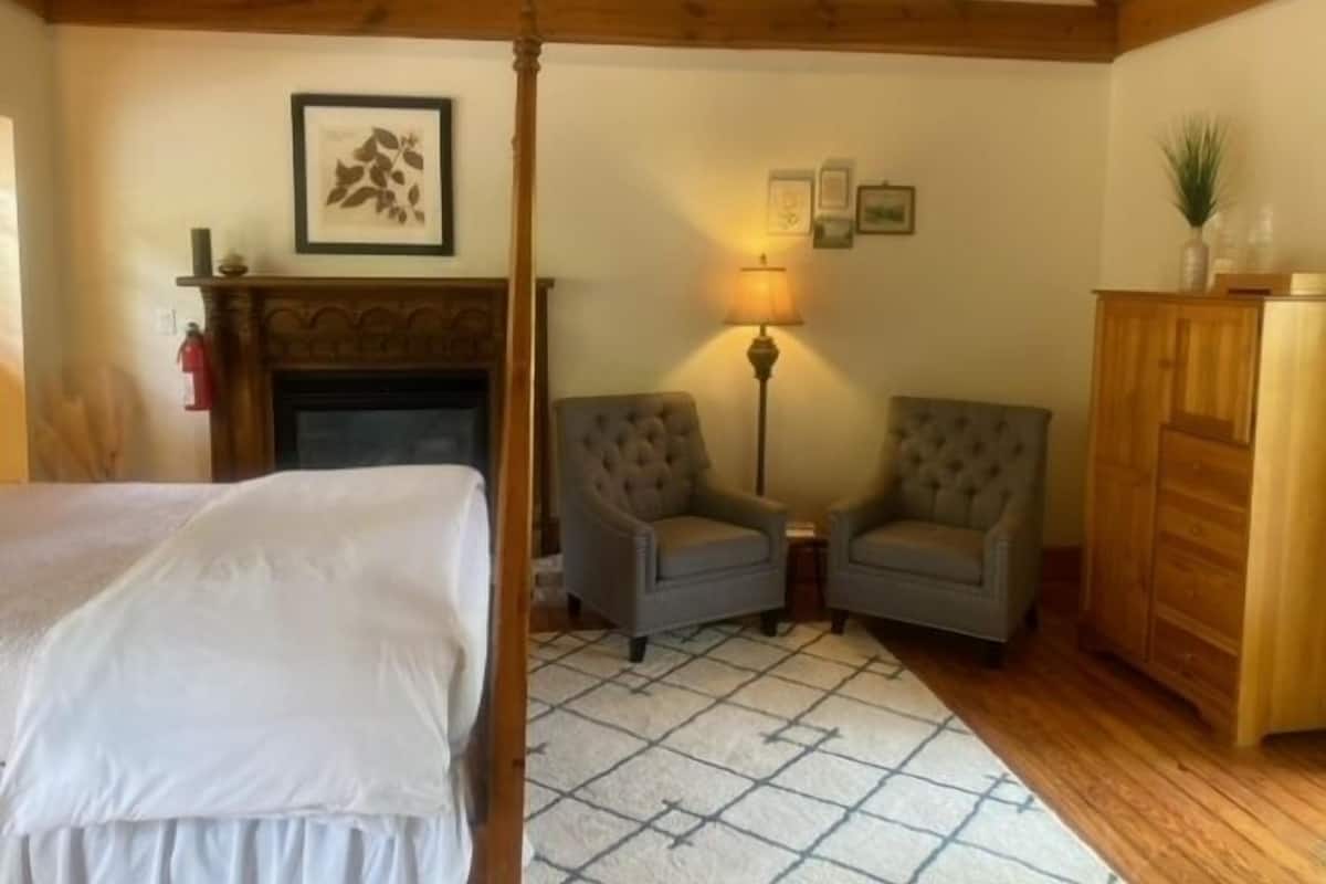 A bedroom with a bed, a fireplace, two armchairs, a floor lamp, and a wooden wardrobe.