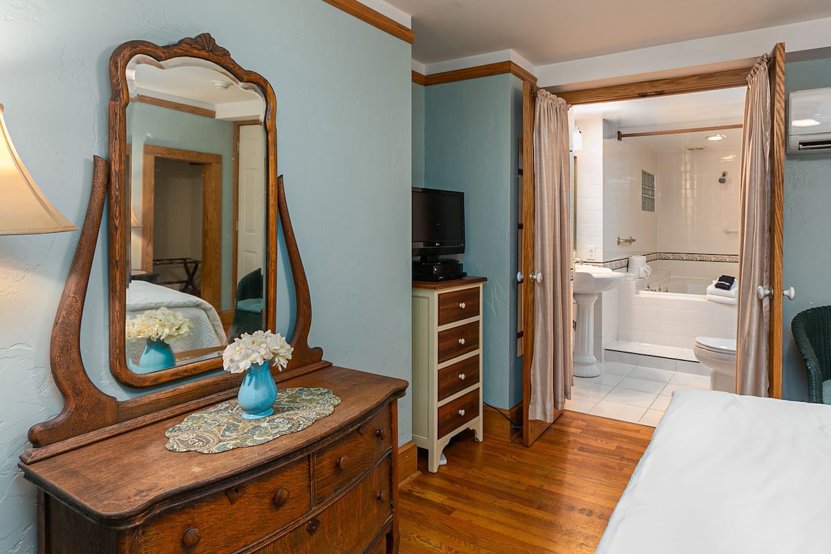 A bedroom with a wooden dresser, a large mirror, a TV, and an open door leading to a bathroom with a bathtub and toilet.