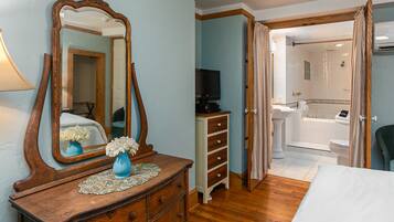 A bedroom with a wooden dresser, a large mirror, a TV, and an open door leading to a bathroom with a bathtub and toilet.