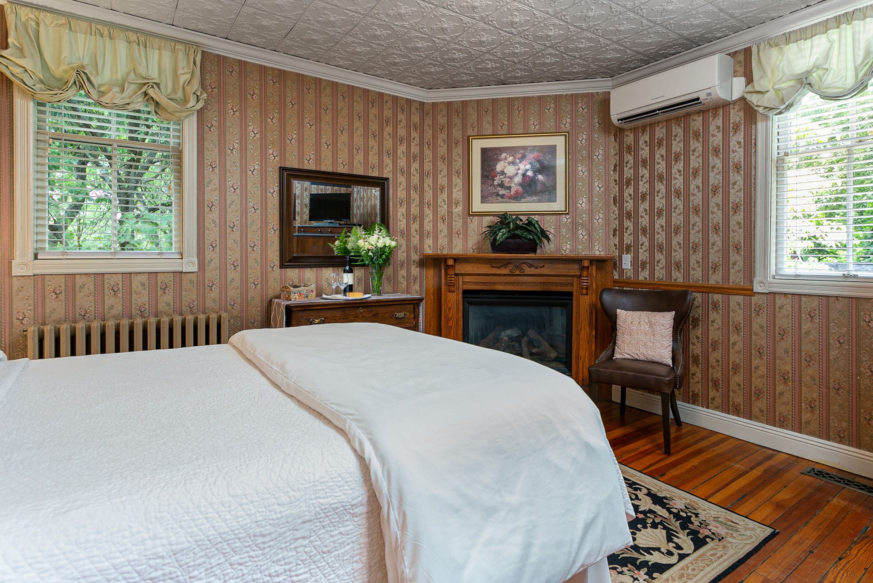 A bedroom with a large bed, a dresser, a mirror, a fireplace, and a window with curtains.