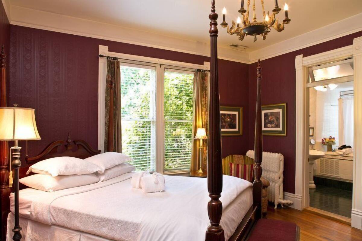 A well-lit bedroom with a four-poster bed, a large window with blinds, and a mirror in the adjacent room.