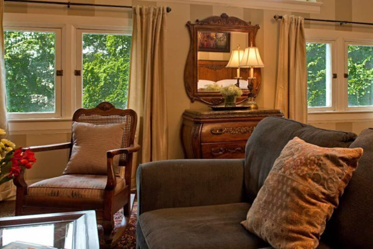 A cozy living room with a brown sofa, a wooden armchair, and a wooden dresser. There is a window with curtains, a mirror, and a vase with flowers on the dresser.