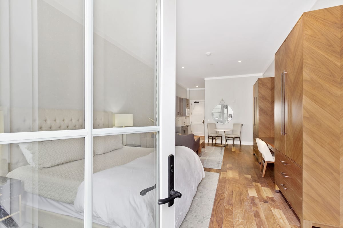 A modern bedroom with a glass door, a wooden wardrobe, a bed with white bedding, and a desk area with a chair.