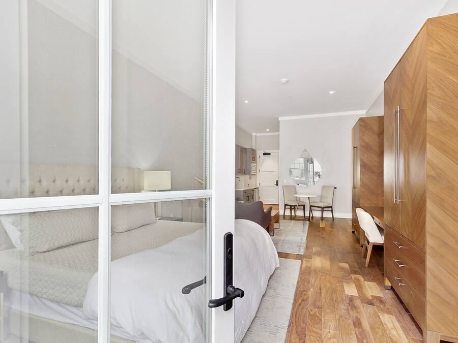 A modern bedroom with a glass door, a wooden wardrobe, a bed with white bedding, and a desk area with a chair.