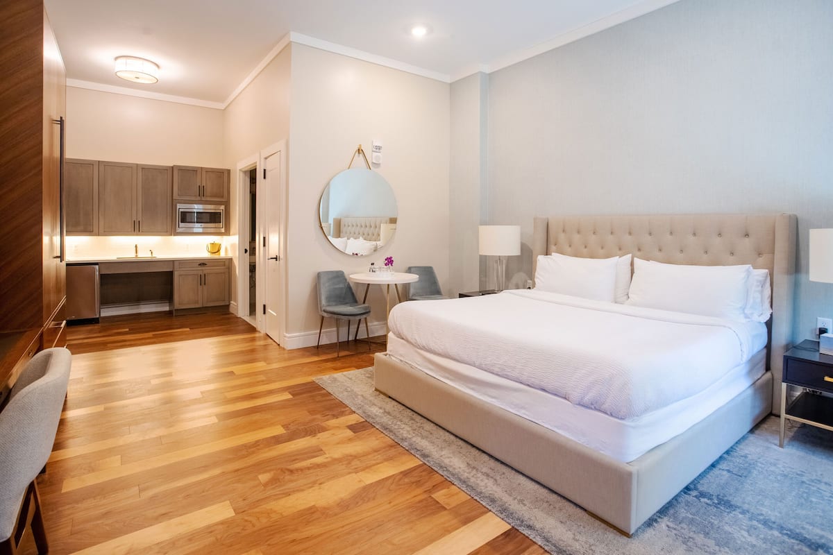 A modern hotel room with a large bed, a small desk, a round mirror, and a kitchenette.