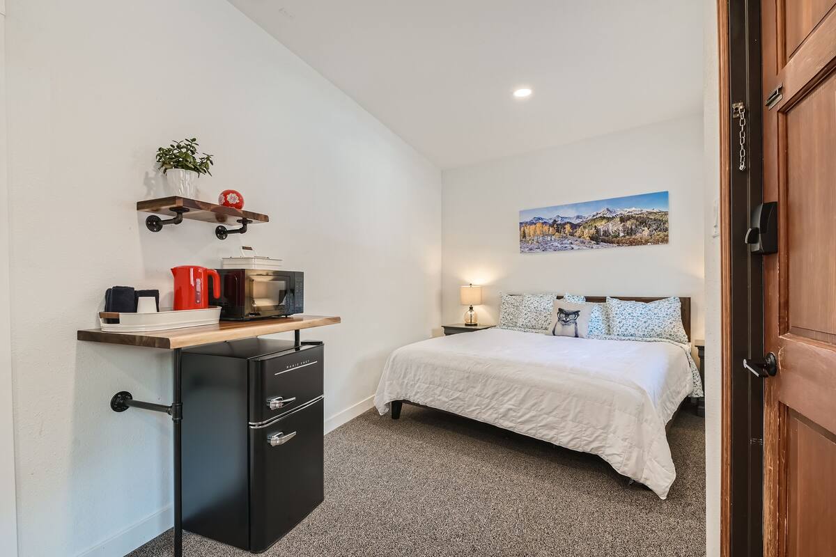 Ein kompaktes Hotelzimmer mit Bett, Schreibtisch mit Kaffeemaschine, kleinem Kühlschrank, Ablagefläche mit Pflanze und roter Tasse sowie einem Bild an der Wand.