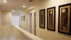 Un couloir avec une rambarde blanche, trois tableaux encadrés représentant des motifs floraux accrochés au mur et une porte au fond.