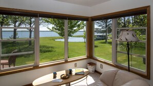 A room with a large bay window overlooking a grassy area and a body of water. There is a table with a lamp, a chair, and some books.