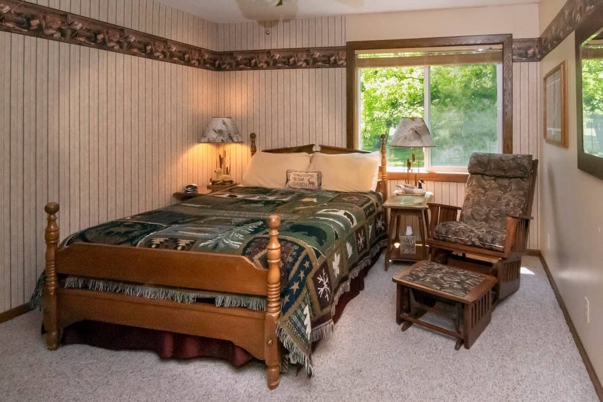 A bedroom with a wooden bed, a chair, a nightstand, and a window with a view of trees.
