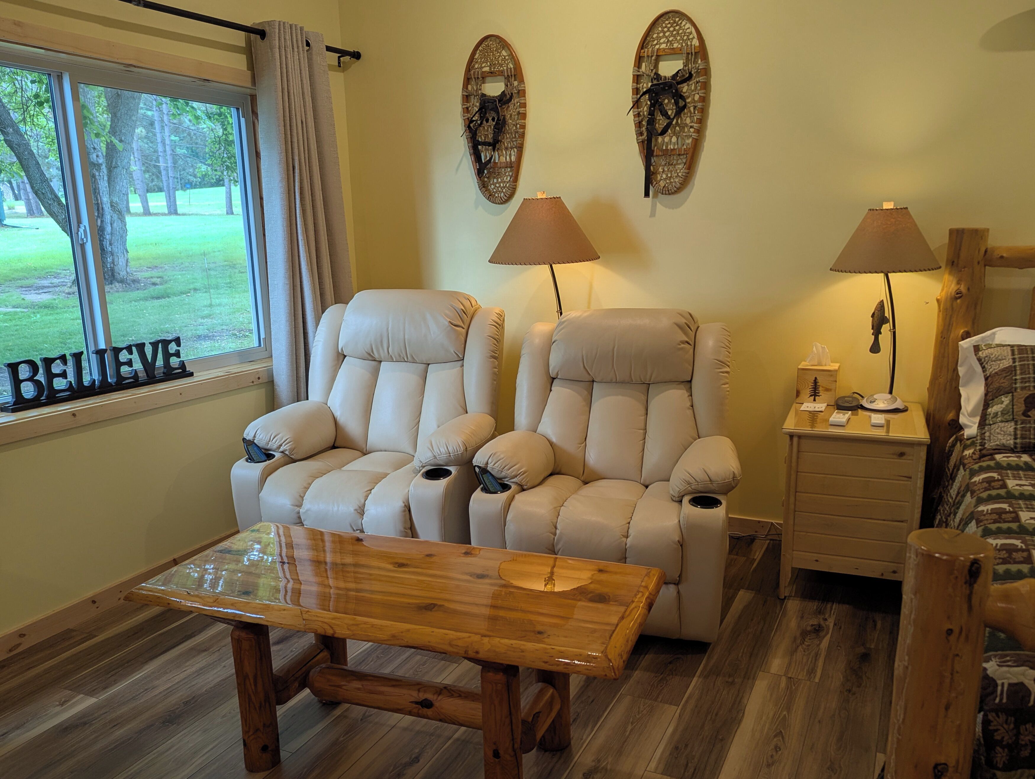 A room with two reclining chairs, a wooden table, a nightstand with a lamp, and a window with a view of trees.