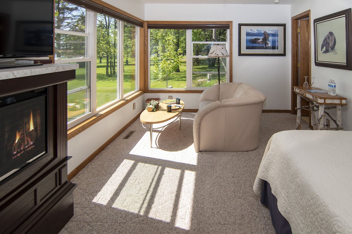 A room with a fireplace, a bed, a chair, a table, and large windows with a view of a green landscape.