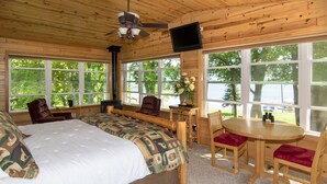 A wooden cabin with a large bed, a dining table, and multiple windows offering views of a lake.