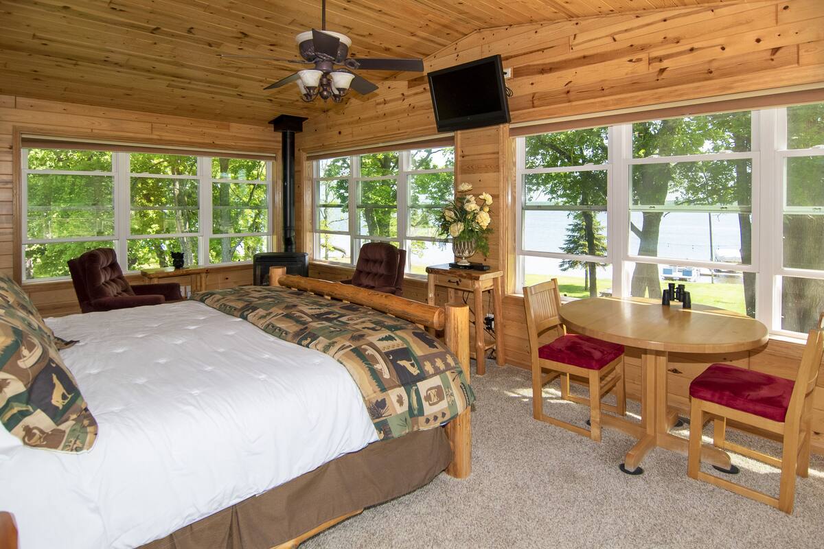 A wooden cabin with a large bed, a dining table, and multiple windows offering views of a lake.