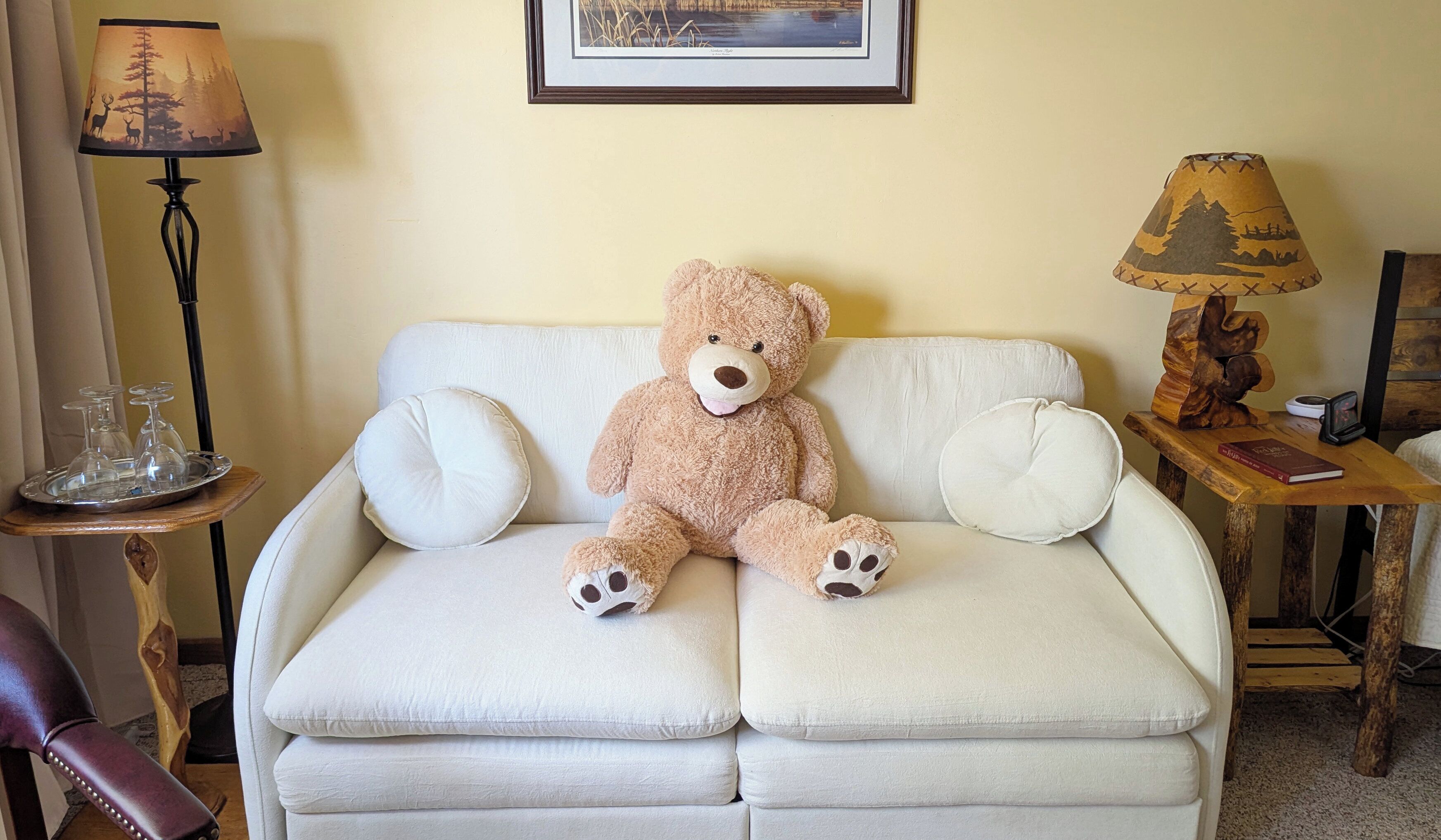Un canapé beige, un grand ours en peluche, une lampe décorée de motifs forestiers et un tableau encadré accroché au mur.
