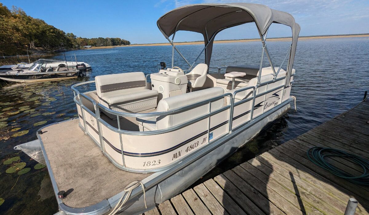 A pontoon boat docked at a wooden pier with a canopy, equipped with seating and a motor.