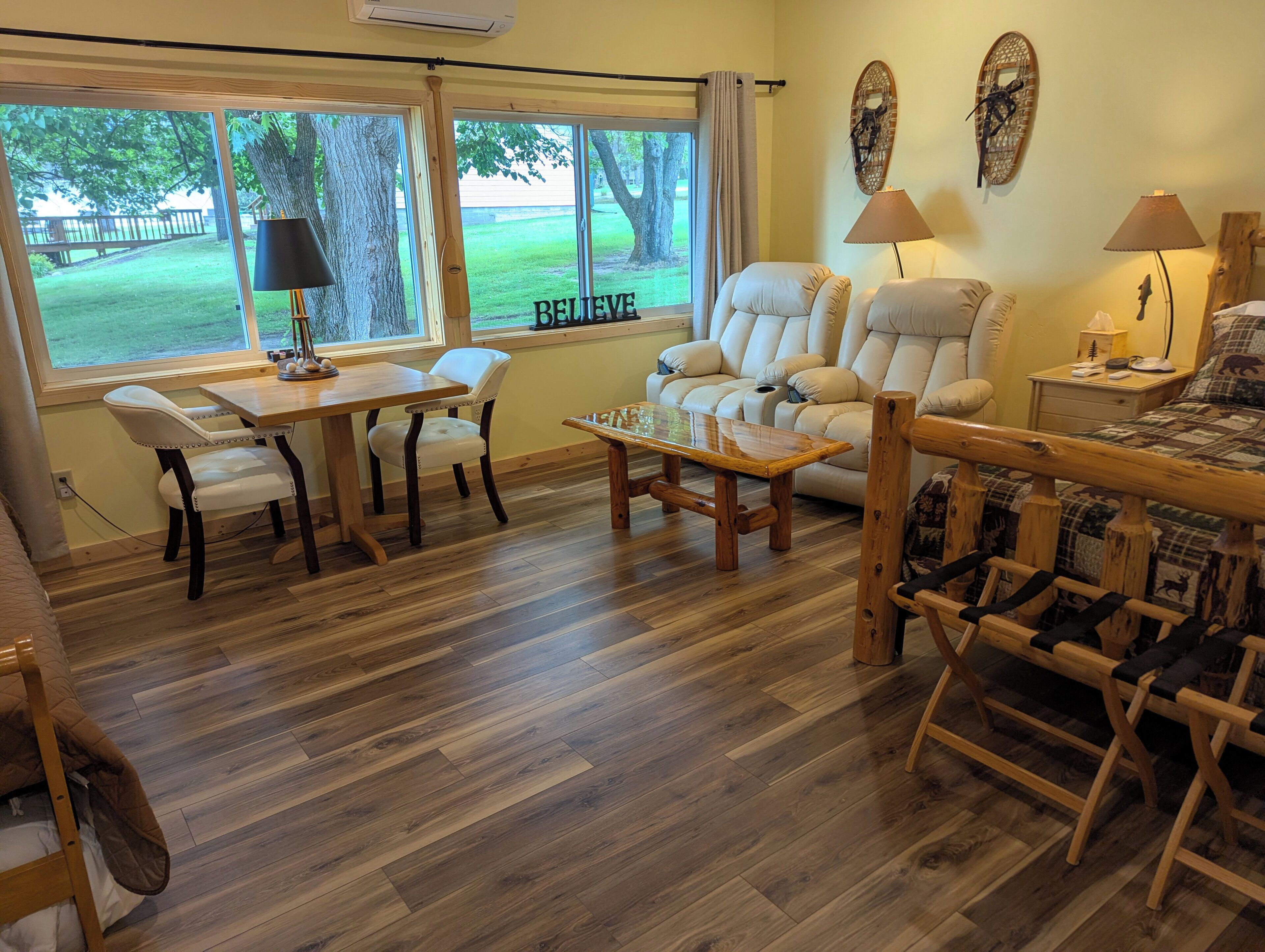 Una sala de estar con piso de madera, un sofá, una mesa de comedor con sillas y vista a un jardín césped a través de grandes ventanales.