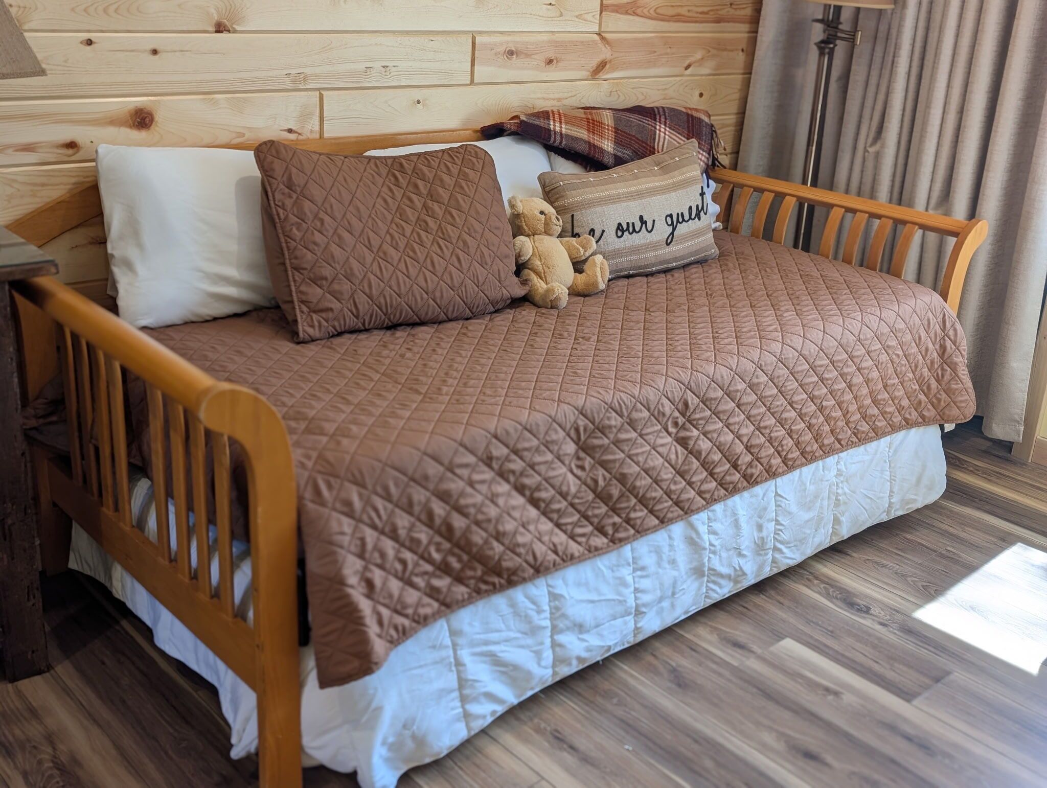 A wooden bed with a quilted brown cover, a white pillow, a plaid pillow, and a teddy bear.
