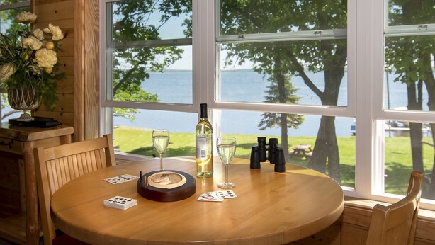 A wooden table with playing cards, a bottle of wine, and glasses, set against a window with a view of a lake and trees.