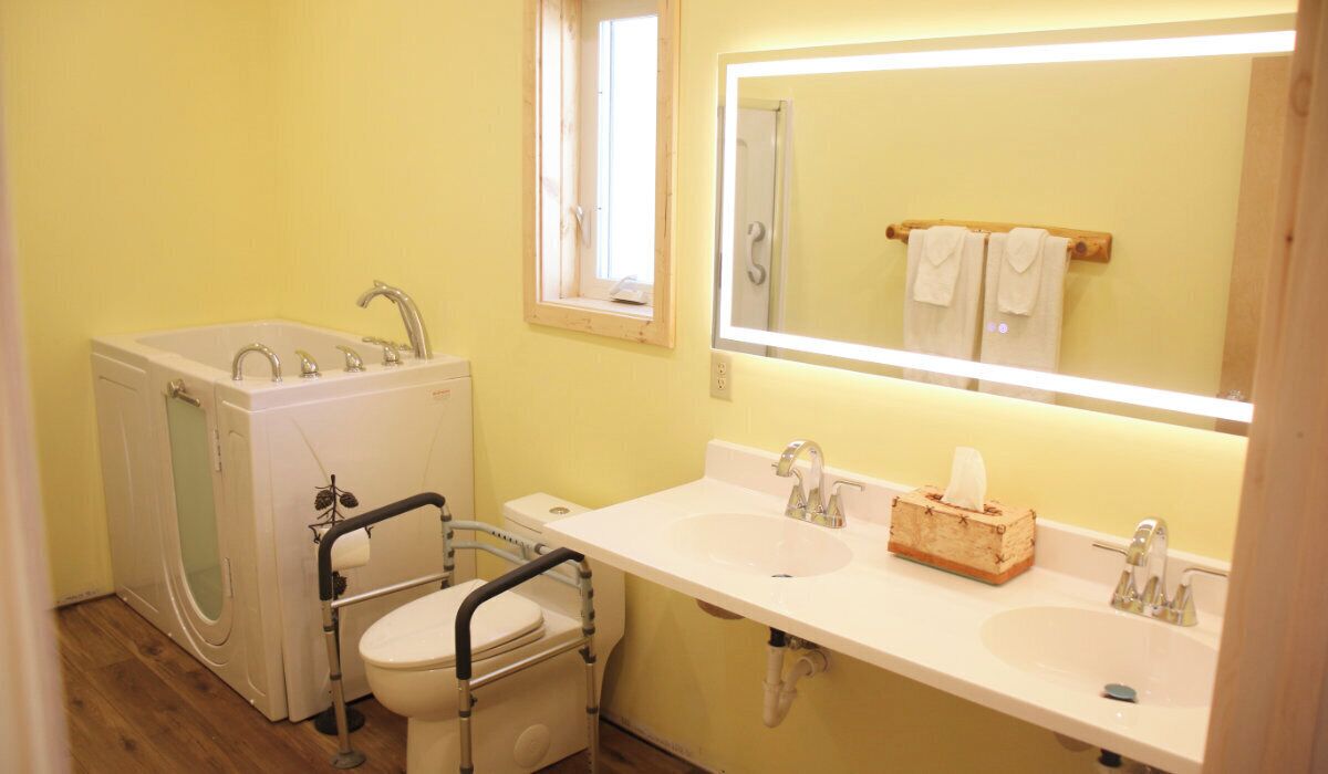 Une salle de bain équipée de toilettes accessibles aux personnes en fauteuil roulant, d’une baignoire et d’un lavabo double avec un miroir au-dessus.