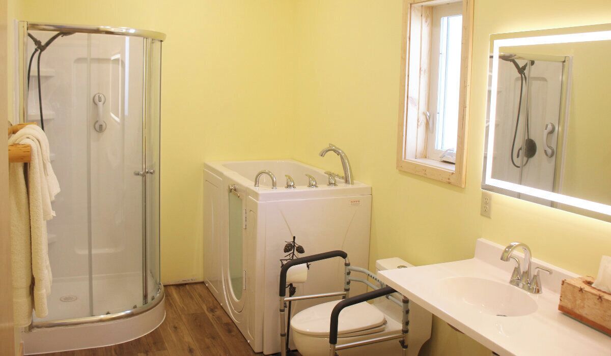 Een badkamer met een glazen douchecabine, een bad, een toilet met een rolstoel toegankelijke zitting en een wastafel met een spiegel erboven.