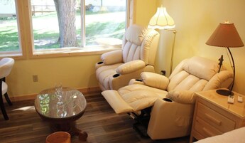 Habitación con sillón de masaje, una mesa de vidrio con copas de vino, una lámpara y vistas al exterior.