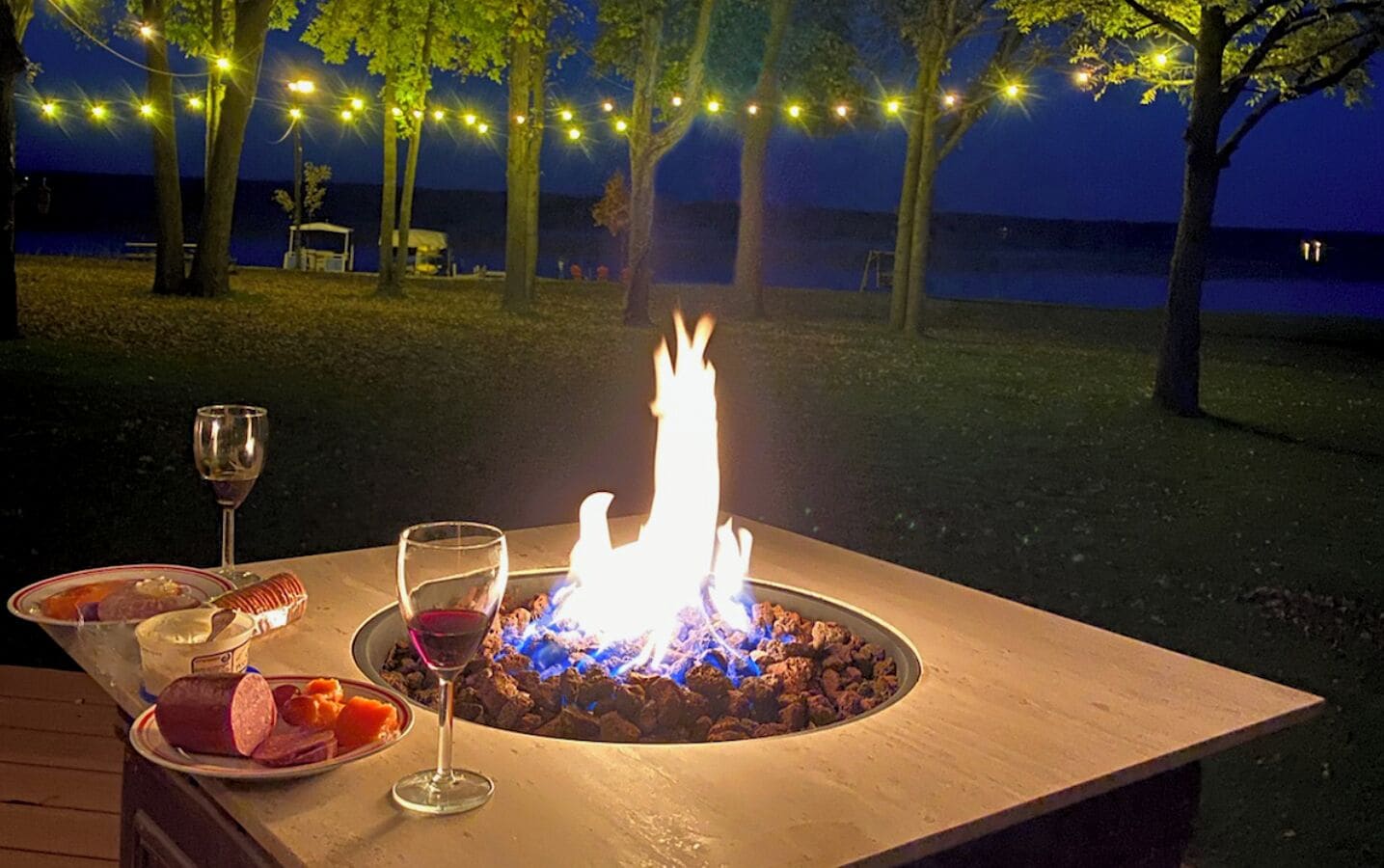 A table with a fire pit, food, and wine glasses set outdoors at night.