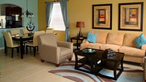 A living room with a sofa, armchair, coffee table, dining table, and chairs. There is a kitchen area with cabinets and a refrigerator. - 6BR 4BA Home in Windsor Hills by CV-2564 (Kissimmee)