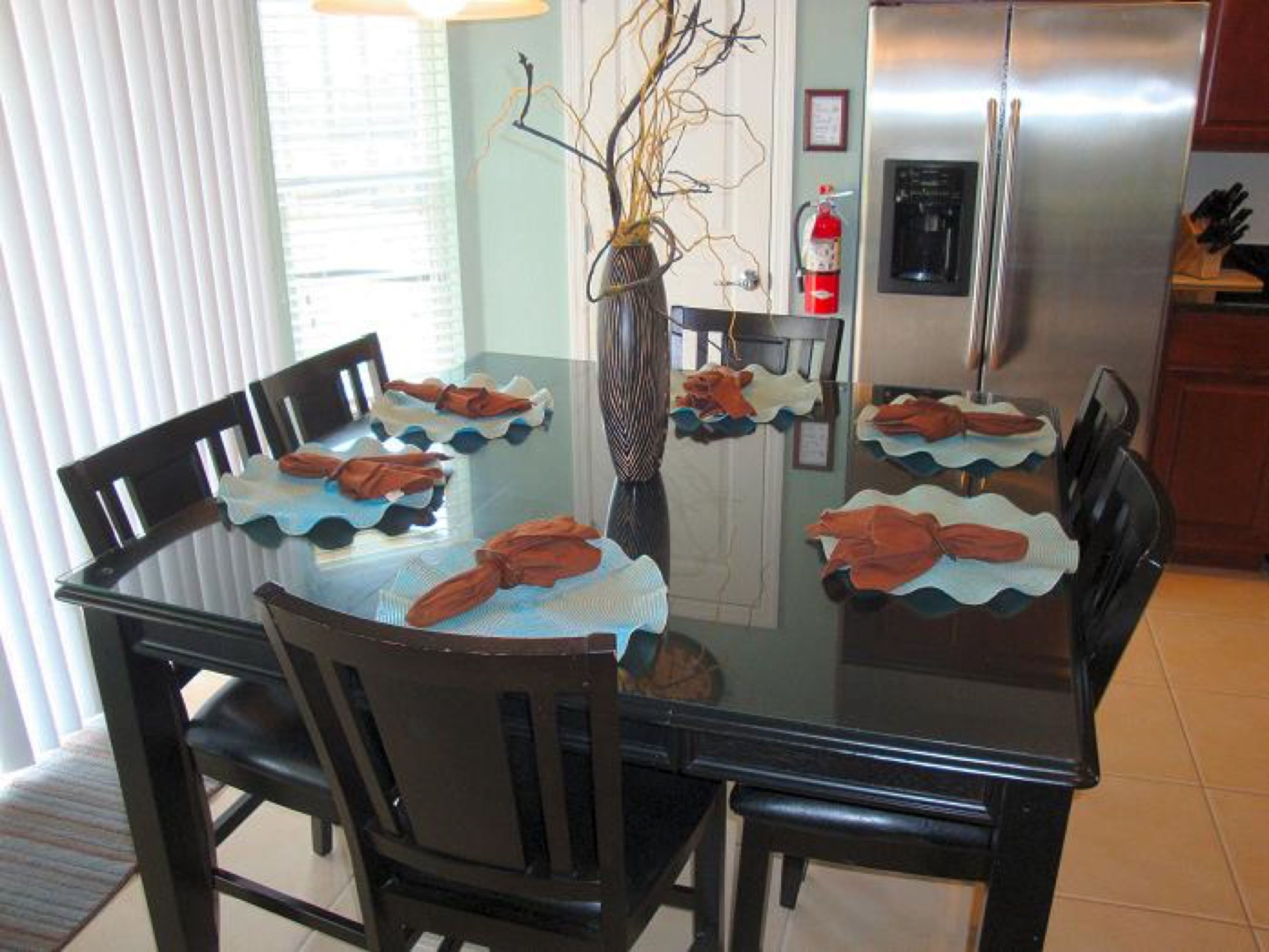 A dining table with chairs, a vase with branches, and a refrigerator in the background.