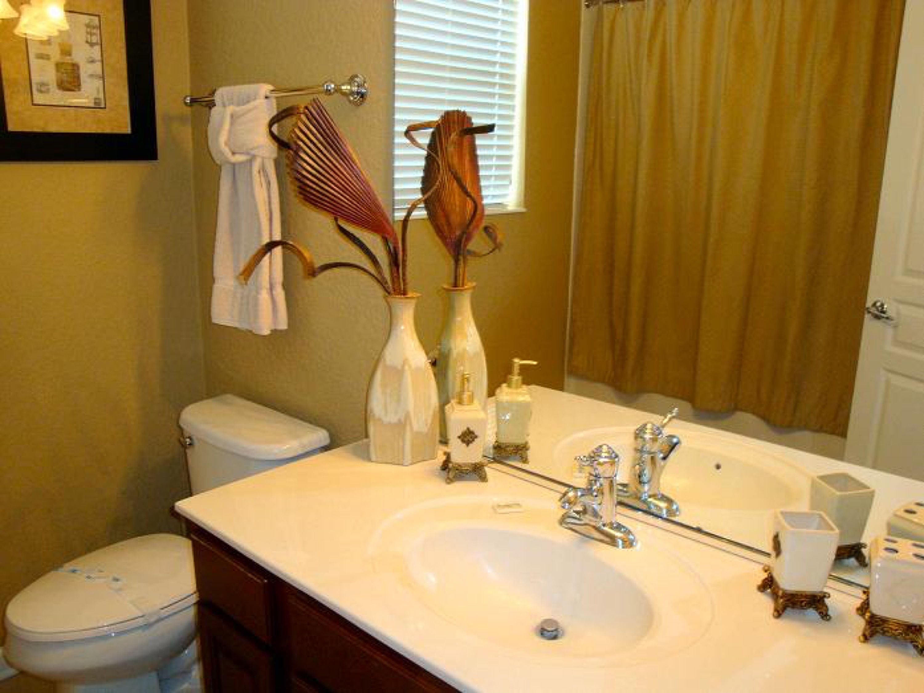 A bathroom with a white sink, two faucets, a toilet, a towel rack, and a vase with dried plants.