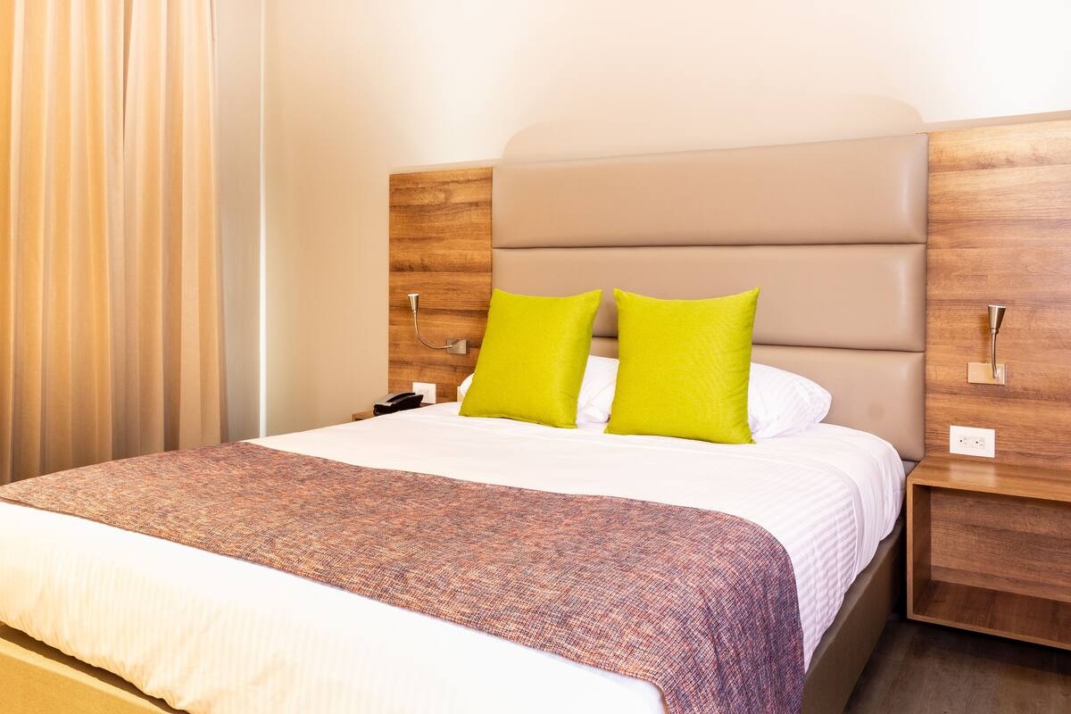 A neatly made bed with a patterned bedspread and two bright pillows, a wooden headboard, and a bedside table with a phone.