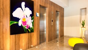 Een moderne lobby met houten lambrisering, een groot schilderij van een orchidee, liften en een felgele stoel.