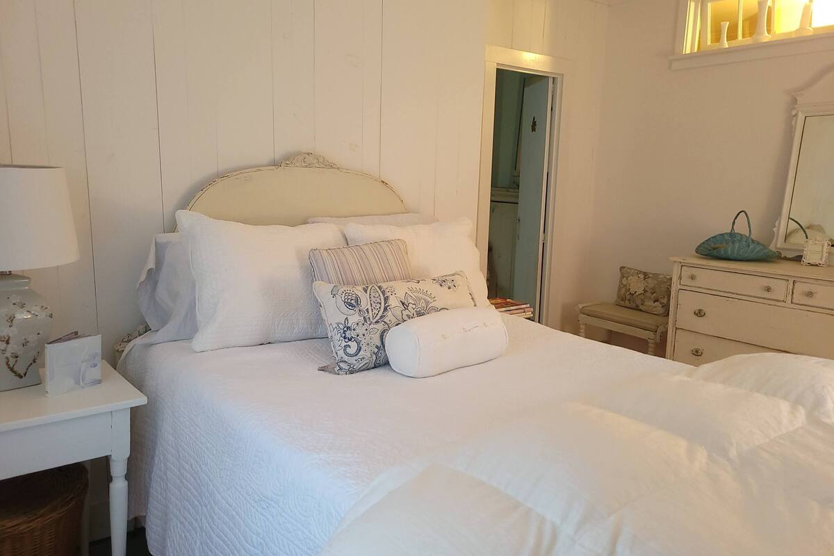 Un lit bien fait, avec une literie blanche et plusieurs oreillers, une table de chevet avec une lampe et une commode avec un miroir.