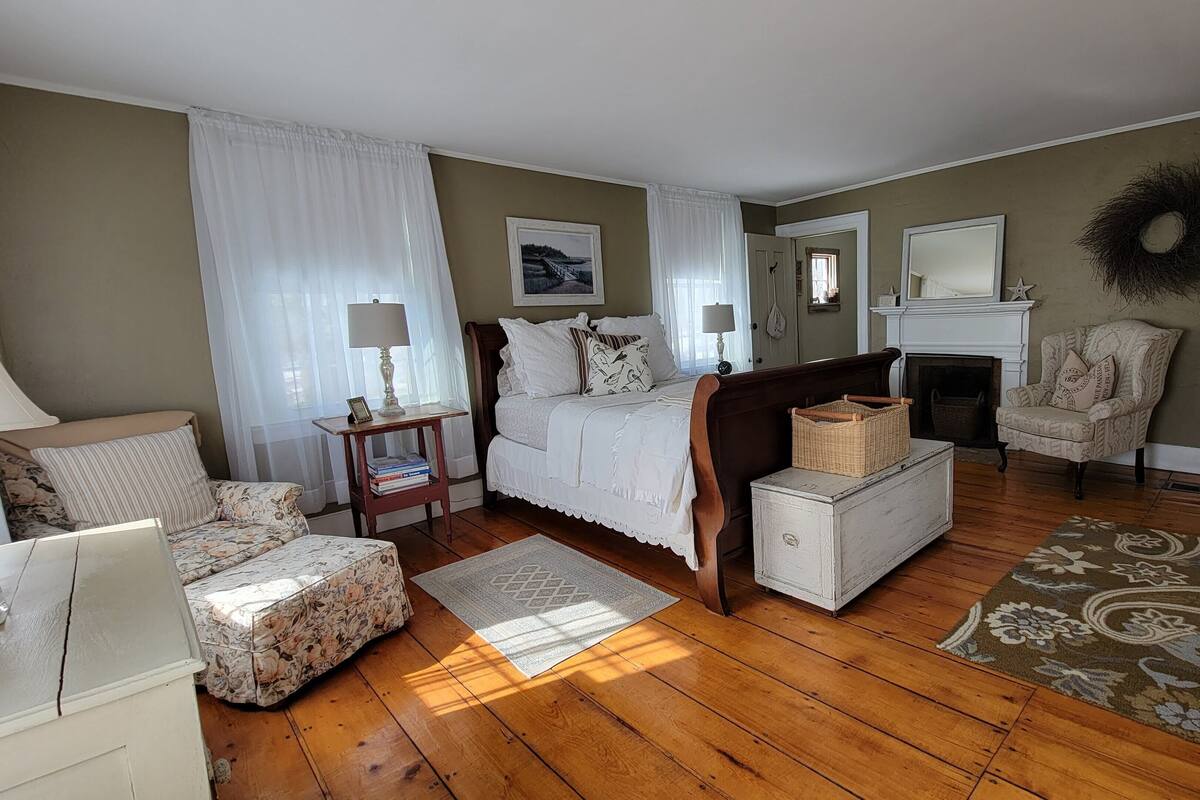 Une chambre avec un plancher en bois, un lit avec une literie blanche, une table de chevet avec une lampe, une chaise, une commode et une cheminée.
