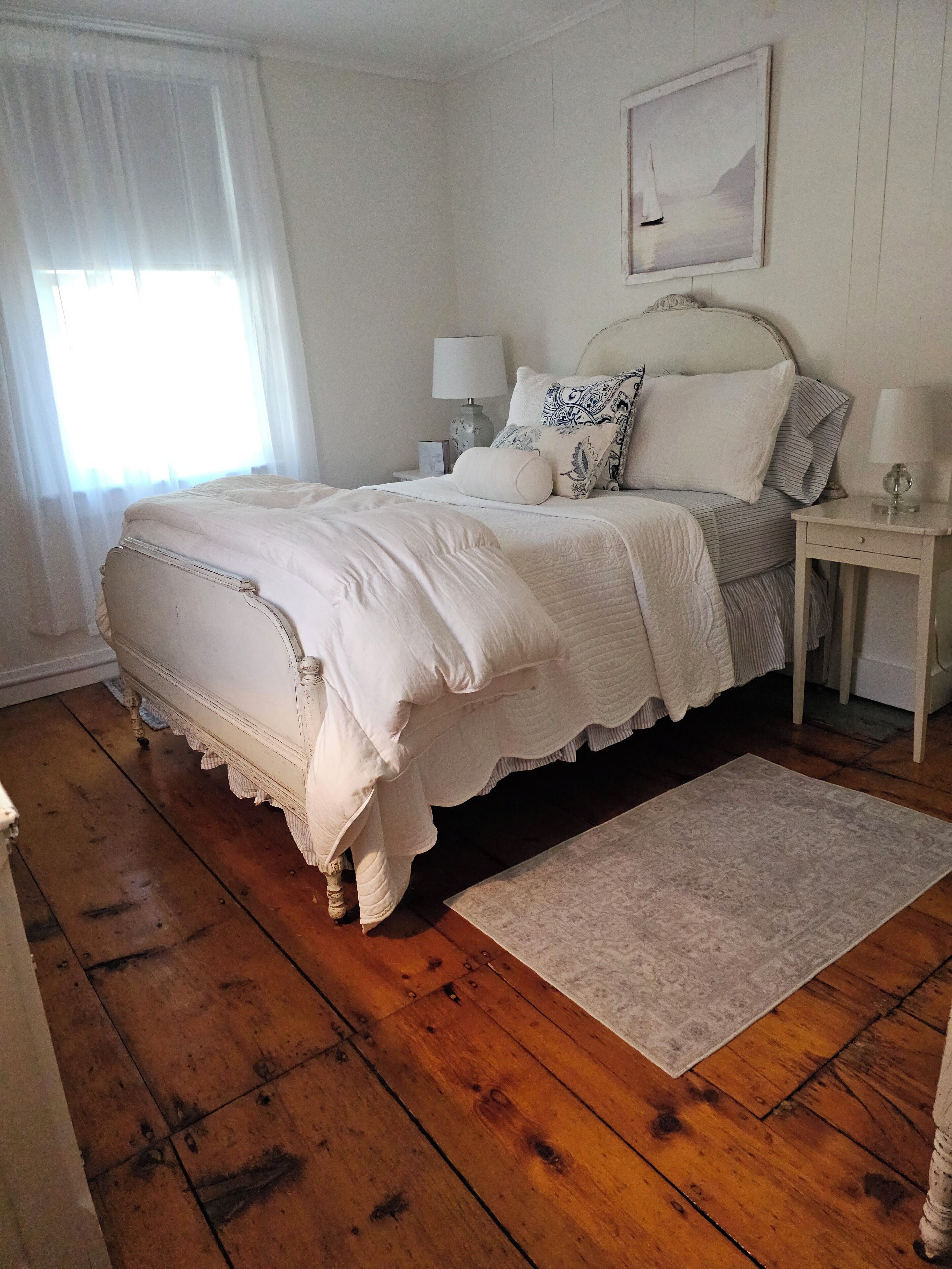 A bedroom with a large bed, a nightstand with a lamp, a window with sheer curtains, and a framed picture on the wall.