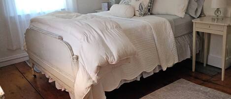 A bedroom with a large bed, a nightstand with a lamp, a window with sheer curtains, and a framed picture on the wall.