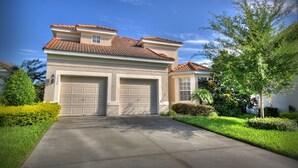 A two-story house with a red-tiled roof, two attached garages, and a well-maintained front yard with a tree and bushes. - 5BR 5BA Home in Windsor HIlls by CV-2622 (Kissimmee)