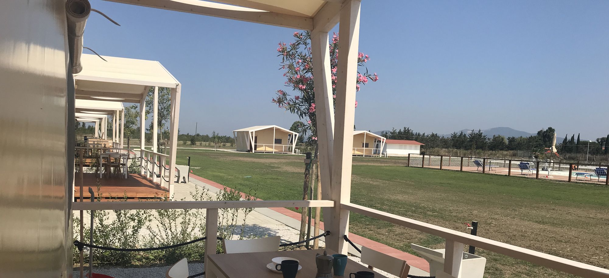 A covered outdoor area with a table and chairs, a view of a grassy field, and a building in the background.