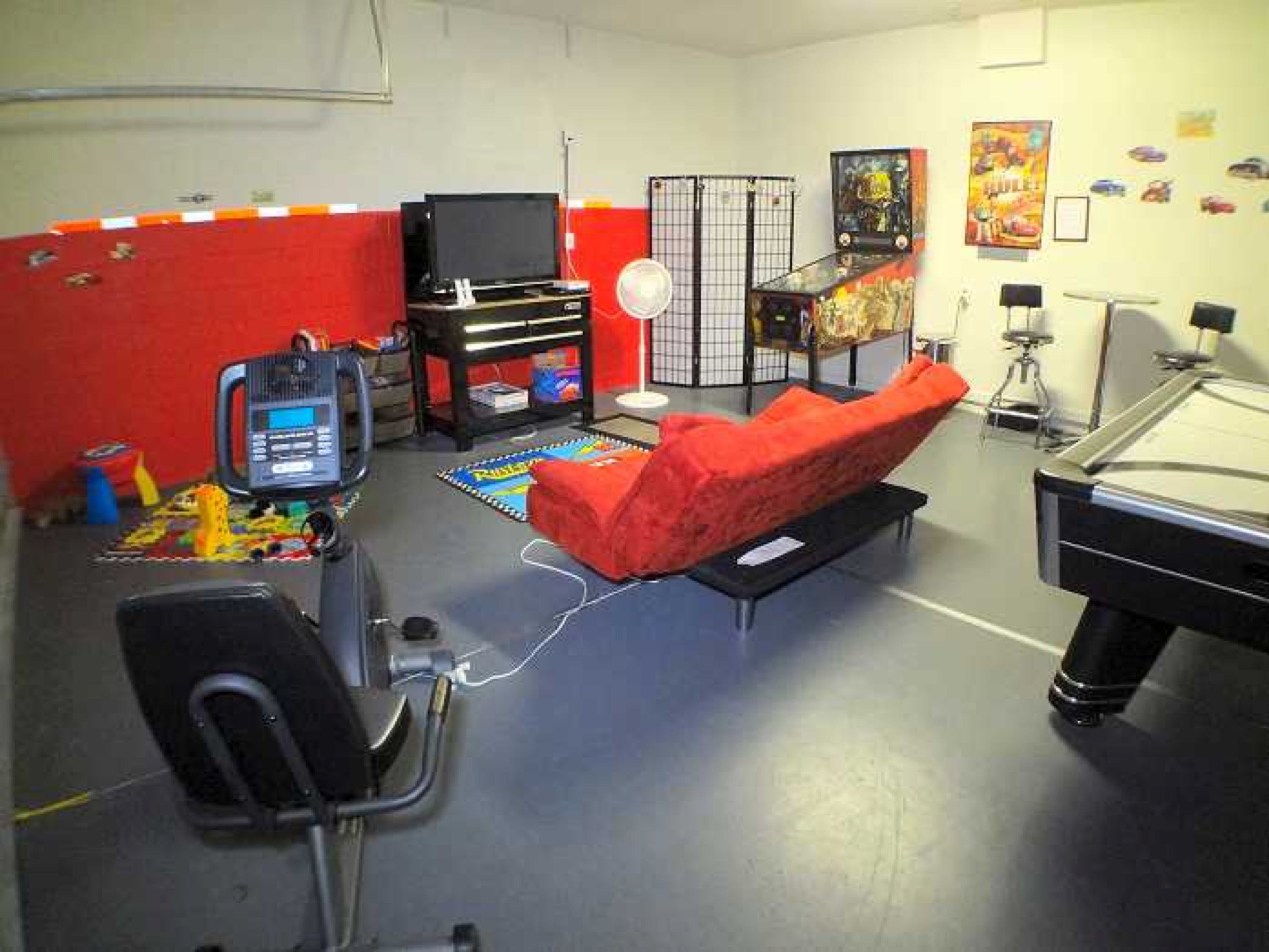 A room with a red couch, a gaming console, a TV, and a pool table.