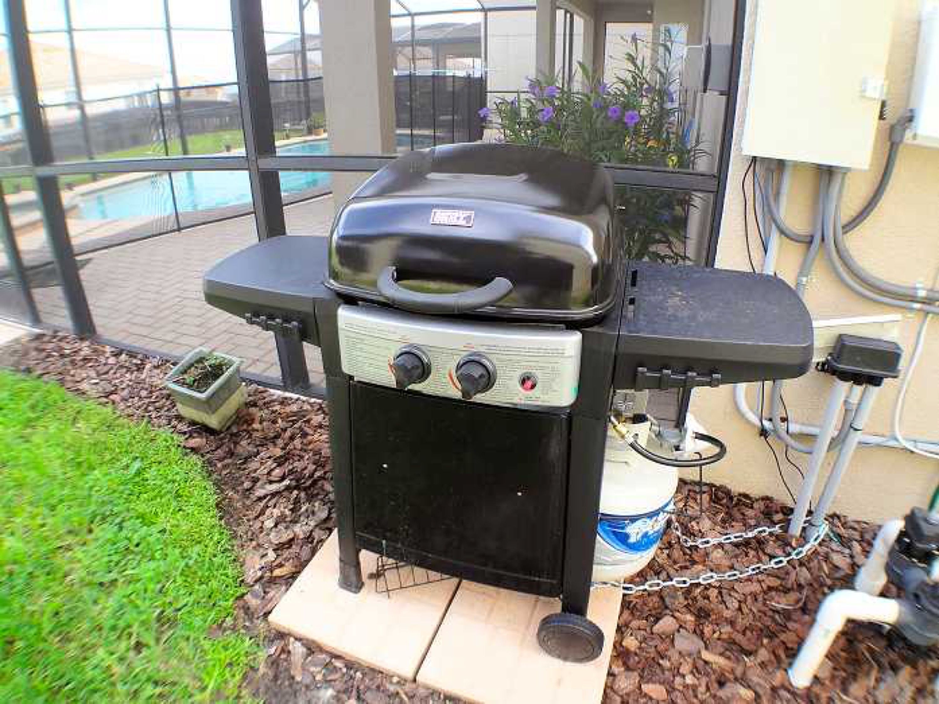 An outdoor grill with controls, a chain for securing it, and a view of a swimming pool in the background.