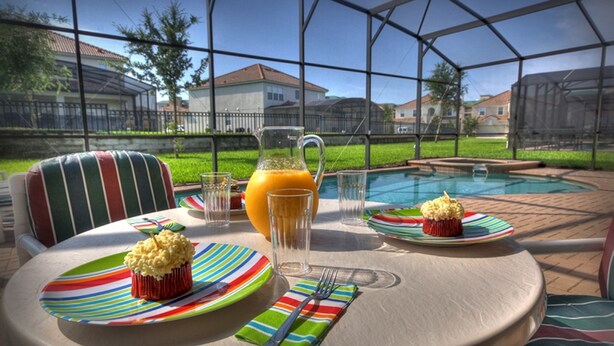 A table set with plates, a pitcher of orange juice, and a cupcake, with a pool and houses in the background. - 6BR 4BA Home in Windsor Hills by CV-2579 (Kissimmee)