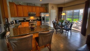 A kitchen with wooden cabinets, a stainless steel refrigerator, a granite countertop, and a dining area with chairs and a table. - 6BR 4BA Home in Windsor Hills by CV-2579 (Kissimmee)
