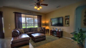 A living room with a sectional sofa, a TV, a ceiling fan, and a large window with curtains. - 6BR 4BA Home in Windsor Hills by CV-2579 (Kissimmee)